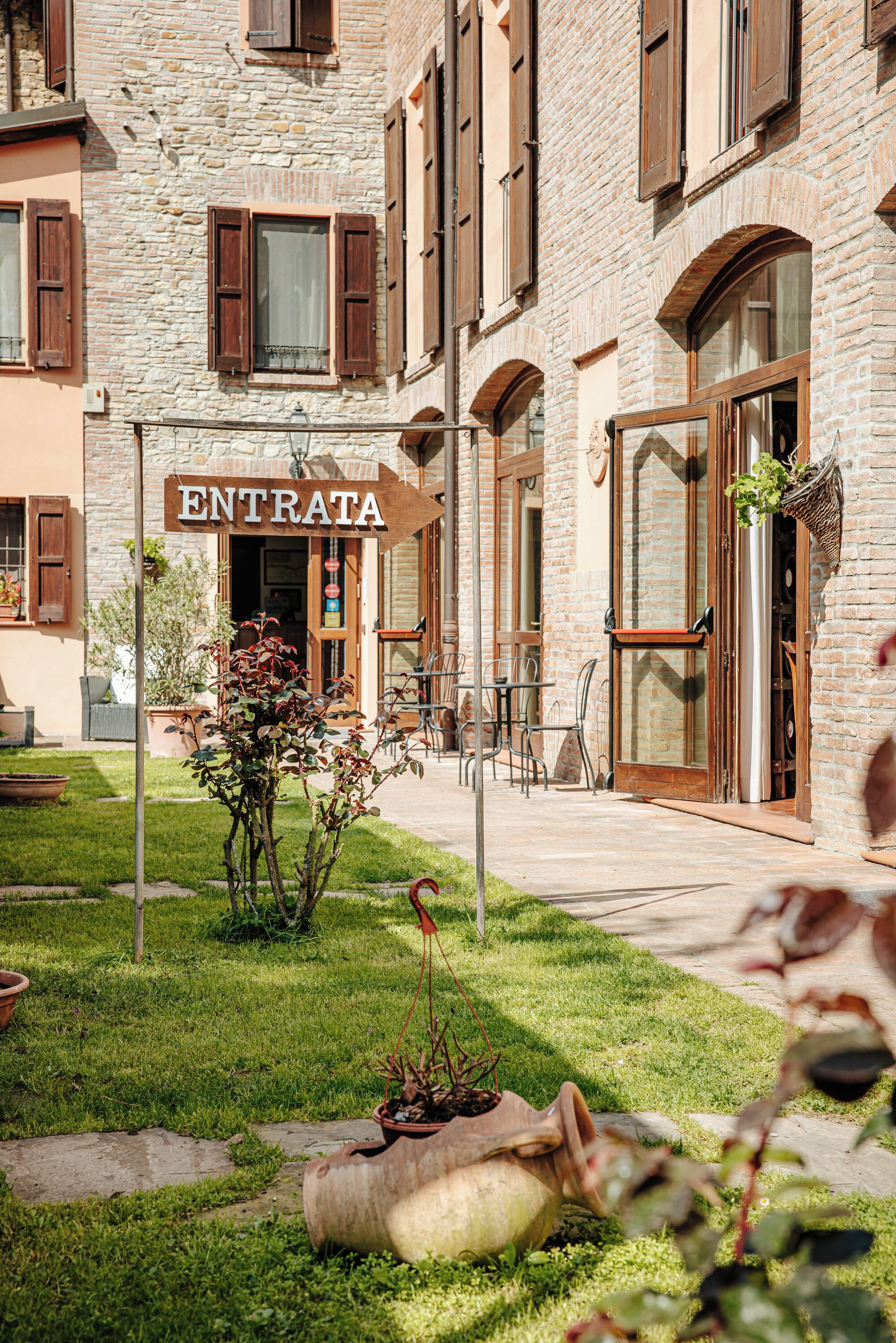 A stone building with a sign reading 'ENTRATA' at the entrance, outdoor seating, and a garden area.