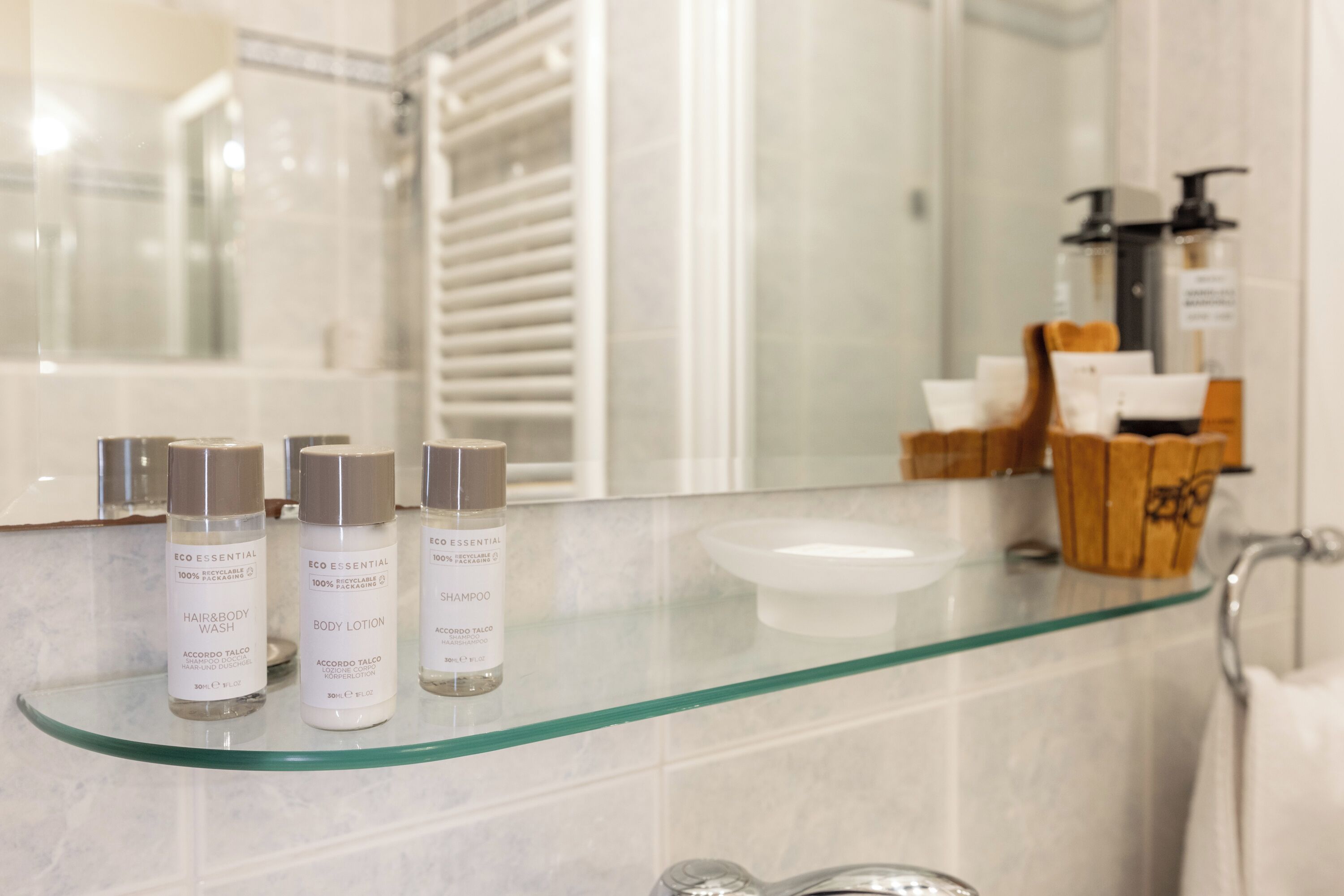 A bathroom with a glass shelf holding three bottles of body lotion and shampoo, a white basin, and a wooden container with toiletries.
