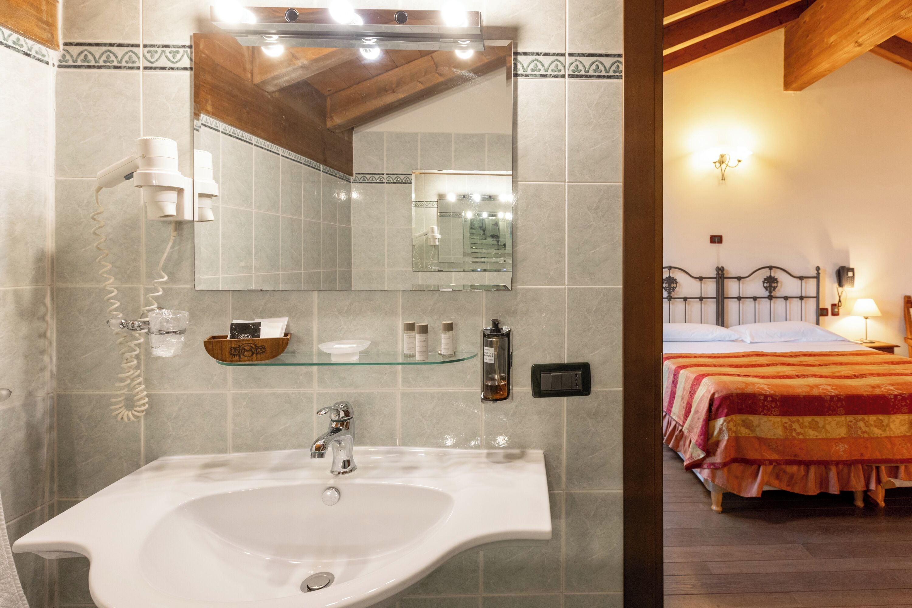 A bathroom with a sink, mirror, and showerhead. There is a bed with a headboard and a bedside table with a lamp.