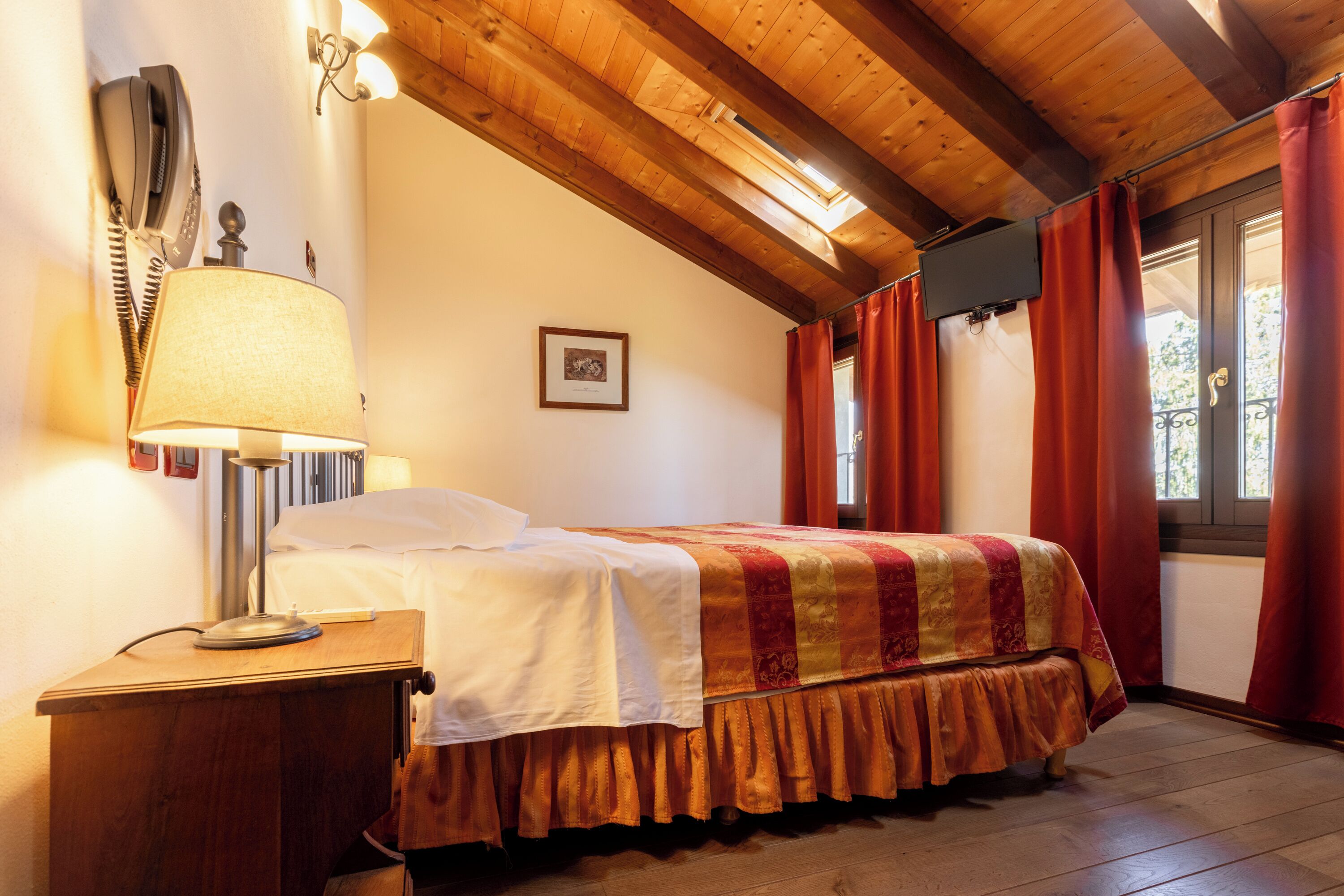 A bedroom with a wooden ceiling, a bed with a patterned bedspread, a bedside lamp, and a window with curtains.