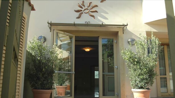 A building entrance with a decorative sun motif above the door, large potted plants on either side, and a clear glass door. - Antella Residence (Bagno a Ripoli)