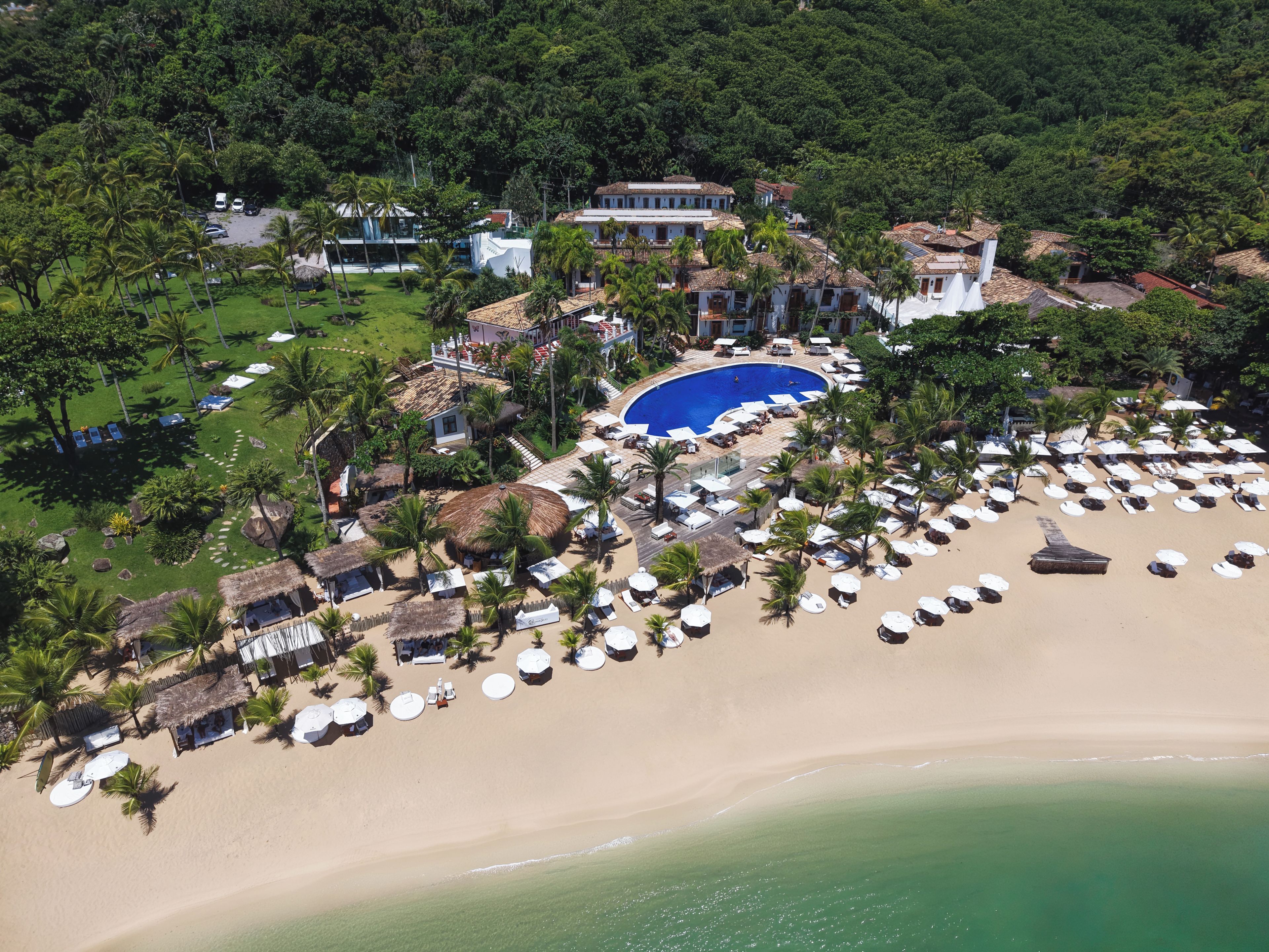 An aerial view of a resort with a large pool, beach area with sun loungers, and lush greenery.