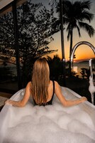 Una persona relajándose en una bañera con vista a la puesta de sol y palmeras.