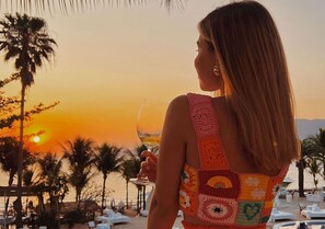 A person holding a glass of white wine, overlooking a beach with palm trees and lounge chairs during sunset. - DPNY Beach Hotel & SPA Ilhabela (Ilhabela)
