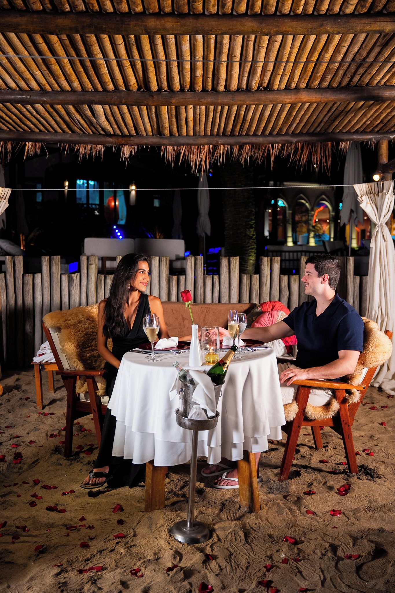 Una pareja cenando en una mesa bajo un techo de paja, rodeada de arena y pétalos de rosa.