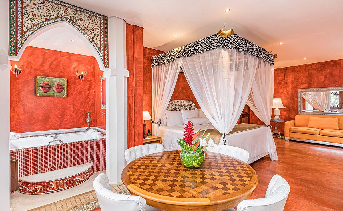 A room with a checkered wooden table, a bed with a canopy, a sofa, a bathtub, and a wall with a red textured finish.