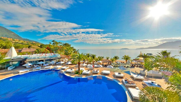 Un resort con una gran piscina, sillas de descanso, sombrillas y vistas al mar y a las montañas.