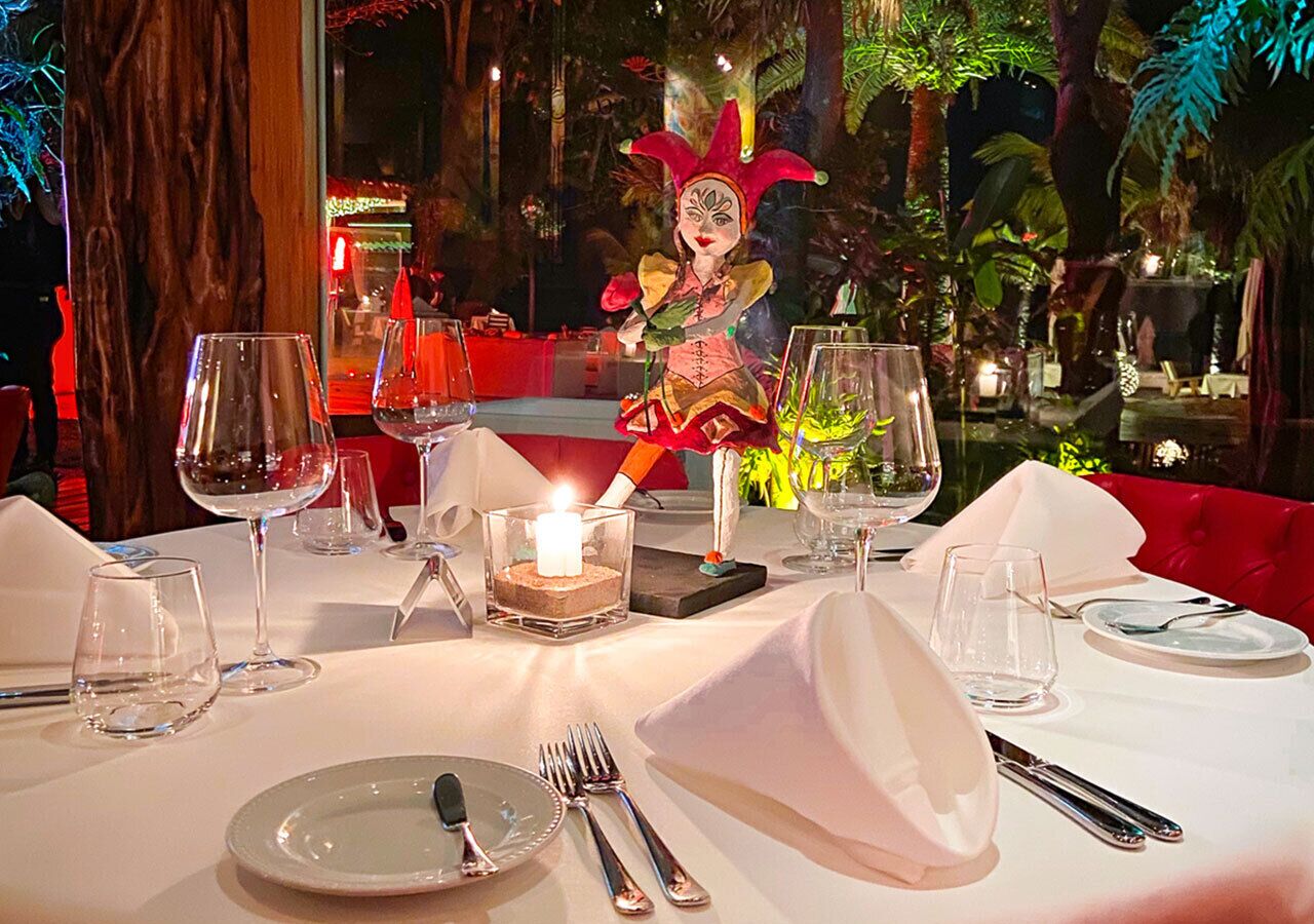 Una mesa de comedor para dos, con un candelabro en el centro, rodeada de plantas tropicales y una figura decorativa.