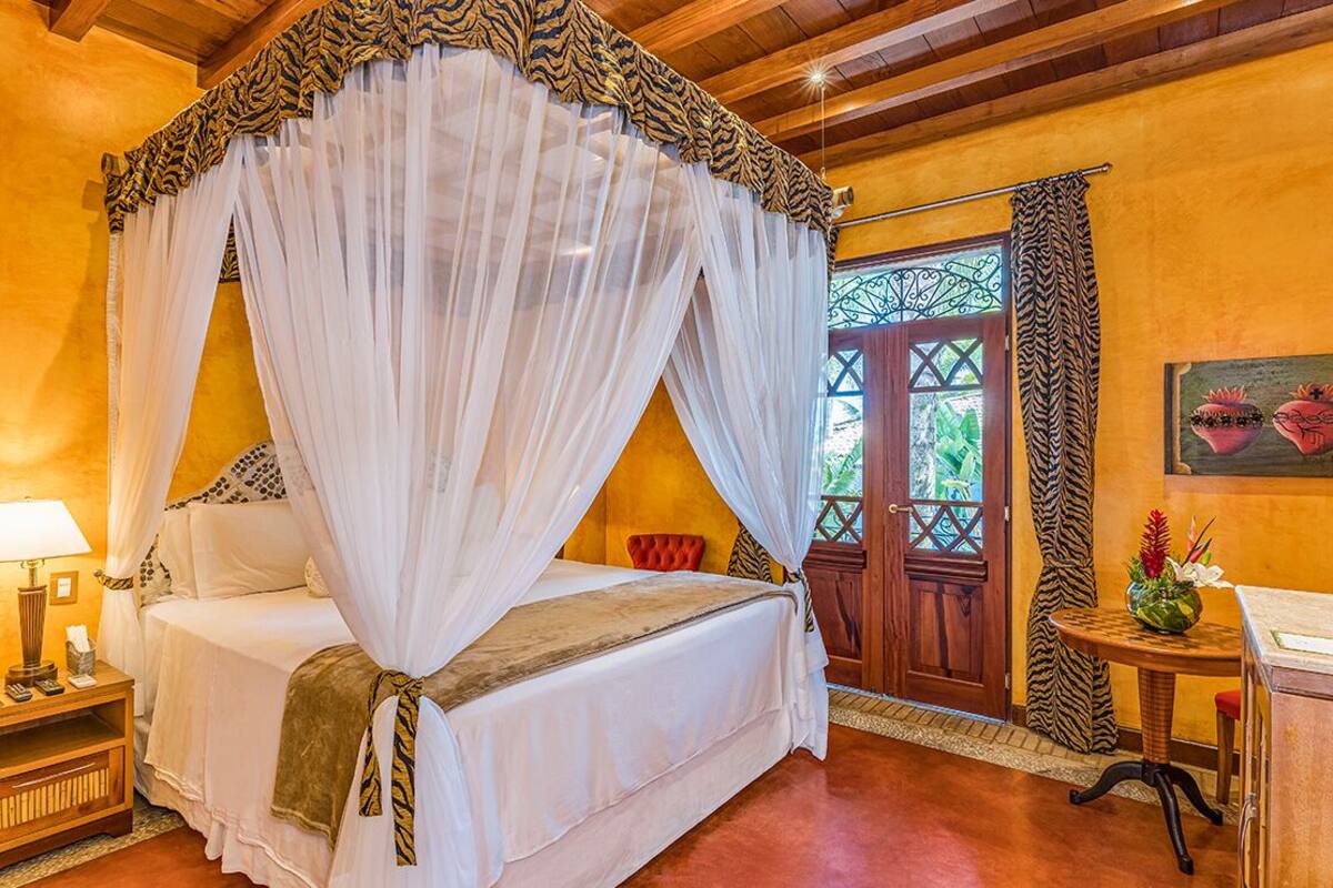 A room with a four-poster bed, a nightstand with a lamp, and a small table with a vase of flowers.