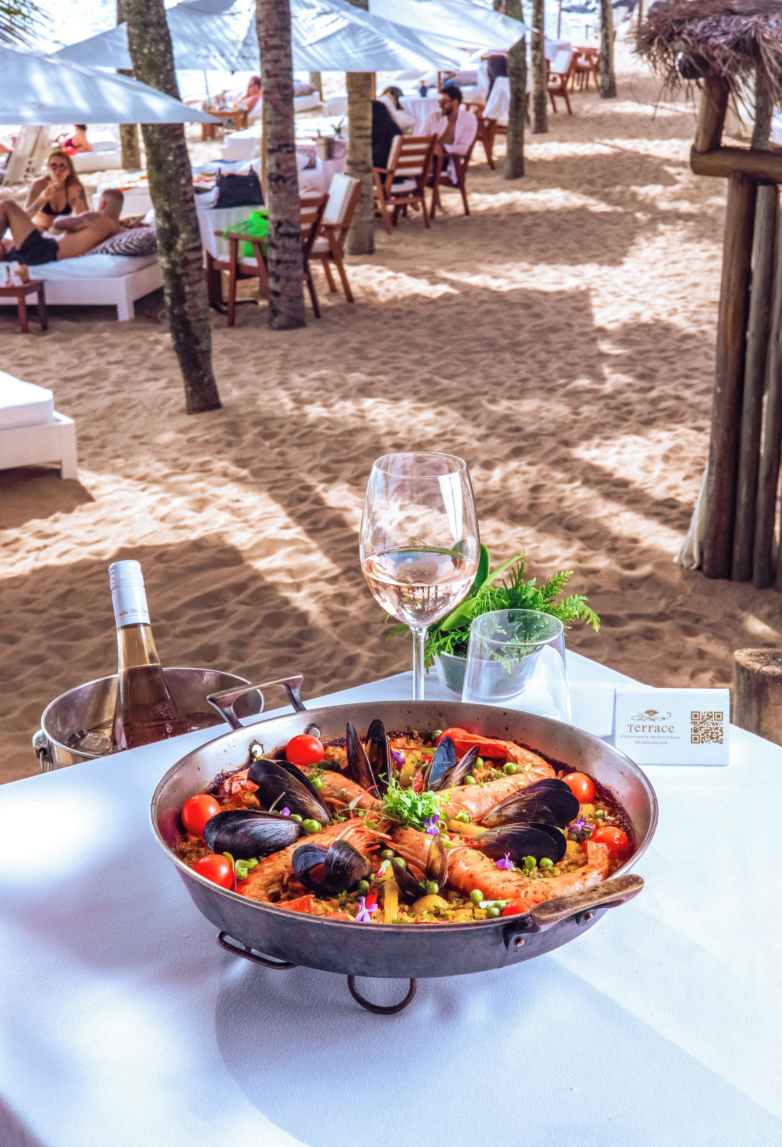 Un plato de paella de mariscos sobre una mesa con una botella de vino y una copa de vino, sobre una playa con sombrillas y sillas de playa al fondo.