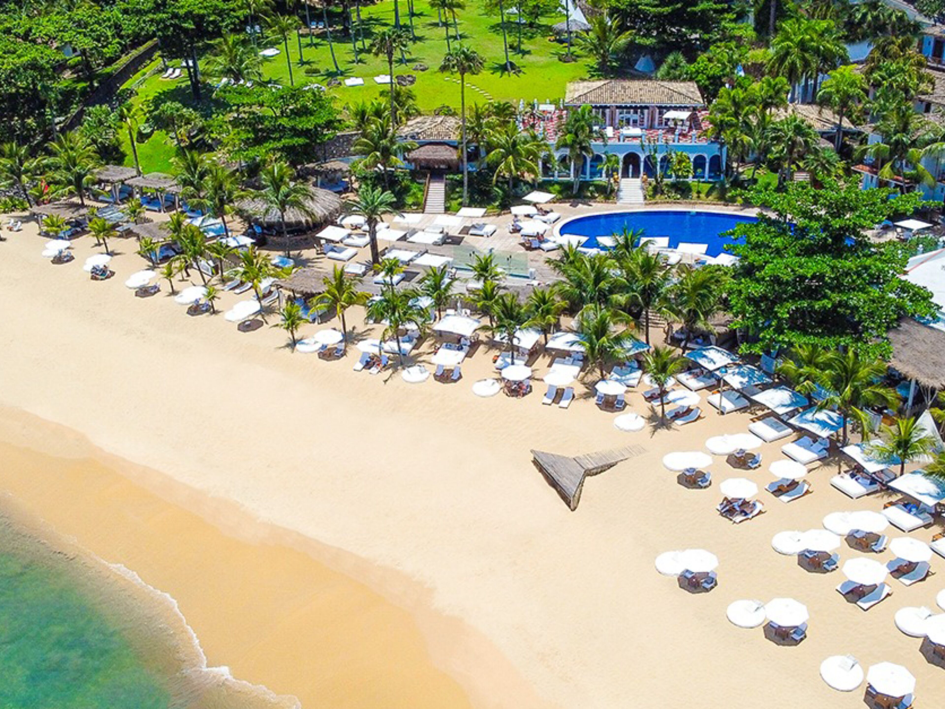 Vista aérea de un complejo turístico en la playa con piscina, tumbonas y palmeras.