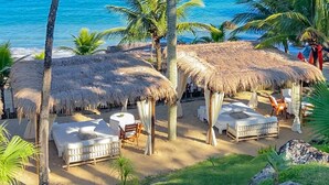 Un resort frente a la playa con cabañas de techo de paja, asientos blancos acolchados y palmeras.
