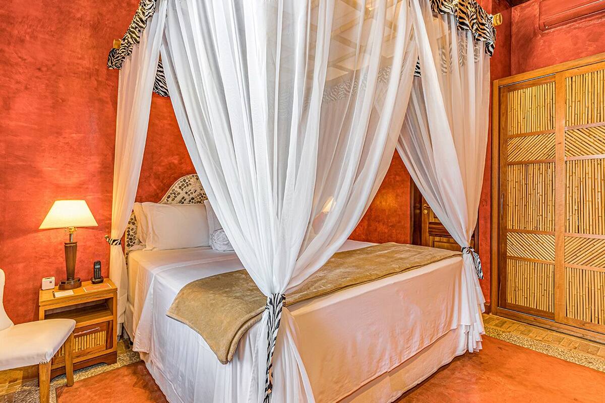 A four-poster bed with white drapes, a bedside table with a lamp, and a wooden wardrobe.