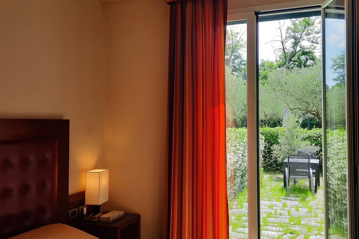 Une chambre d’hôtel avec un lit, une table de chevet avec une lampe et une vue sur un jardin, visible à travers une porte-fenêtre coulissante.