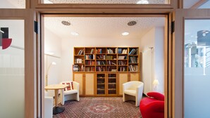 A modern library with a large bookshelf, two white armchairs, a red bean bag, and a patterned rug. - Hotel Am Froschbächel (Bühl)