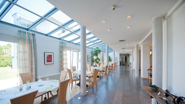 A spacious indoor area with a long corridor, large windows, and several tables and chairs arranged for dining. - Hotel Am Froschbächel (Bühl)