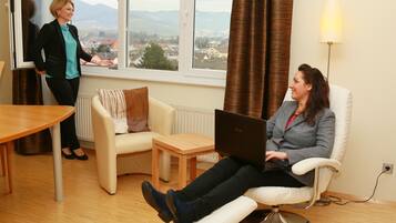 A room with a view of a cityscape through large windows, a white sofa, a wooden table, and a laptop on a reclining chair.