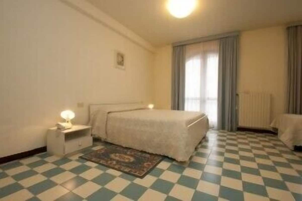 Une chambre avec un sol à carreaux, un lit recouvert d’une couverture blanche, une table de chevet avec une lampe et une fenêtre avec des rideaux.