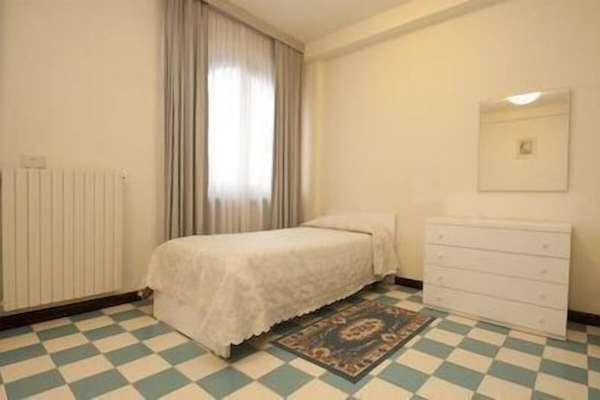 Une chambre à coucher avec un lit, une commode, un radiateur et une fenêtre avec des rideaux.