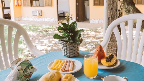 Une table dressée avec des aliments pour le petit-déjeuner, notamment des petits pains, un fruit et un verre de jus d’orange, avec en arrière-plan la vue d’un bâtiment et d’un arbre.