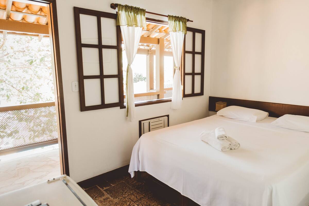A hotel room with a bed, white curtains, a window with a view, and a wooden headboard.