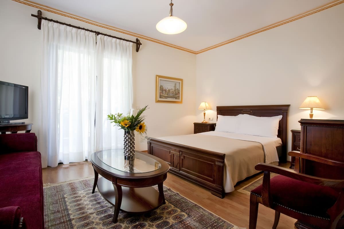 Habitación de hotel con una cama, dos lámparas de noche, un televisor, un sillón rojo, una mesa redonda con un florero y una alfombra estampada.