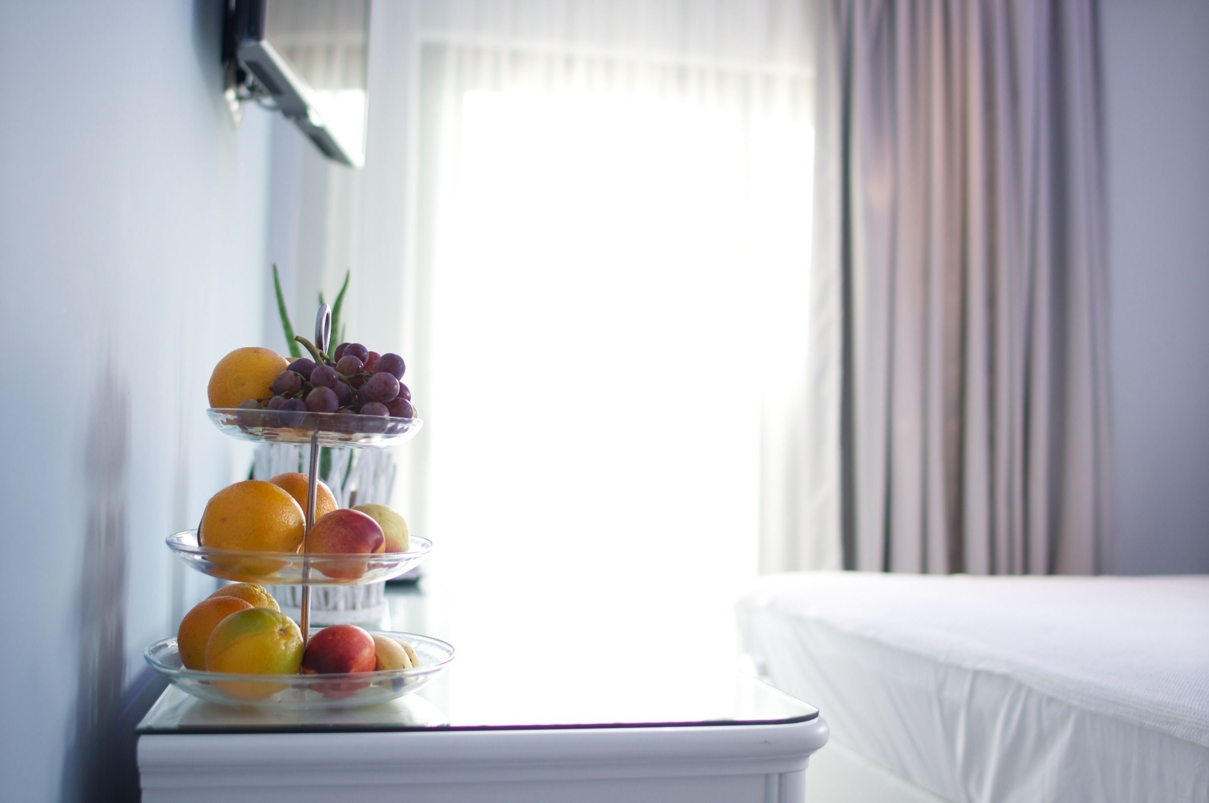 Un puesto de frutas de tres niveles con naranjas, manzanas y uvas sobre una mesa blanca cerca de una ventana con cortinas.