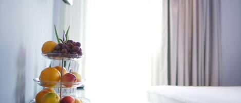 A three-tiered fruit stand with oranges, apples, and grapes on a white table near a window with curtains.