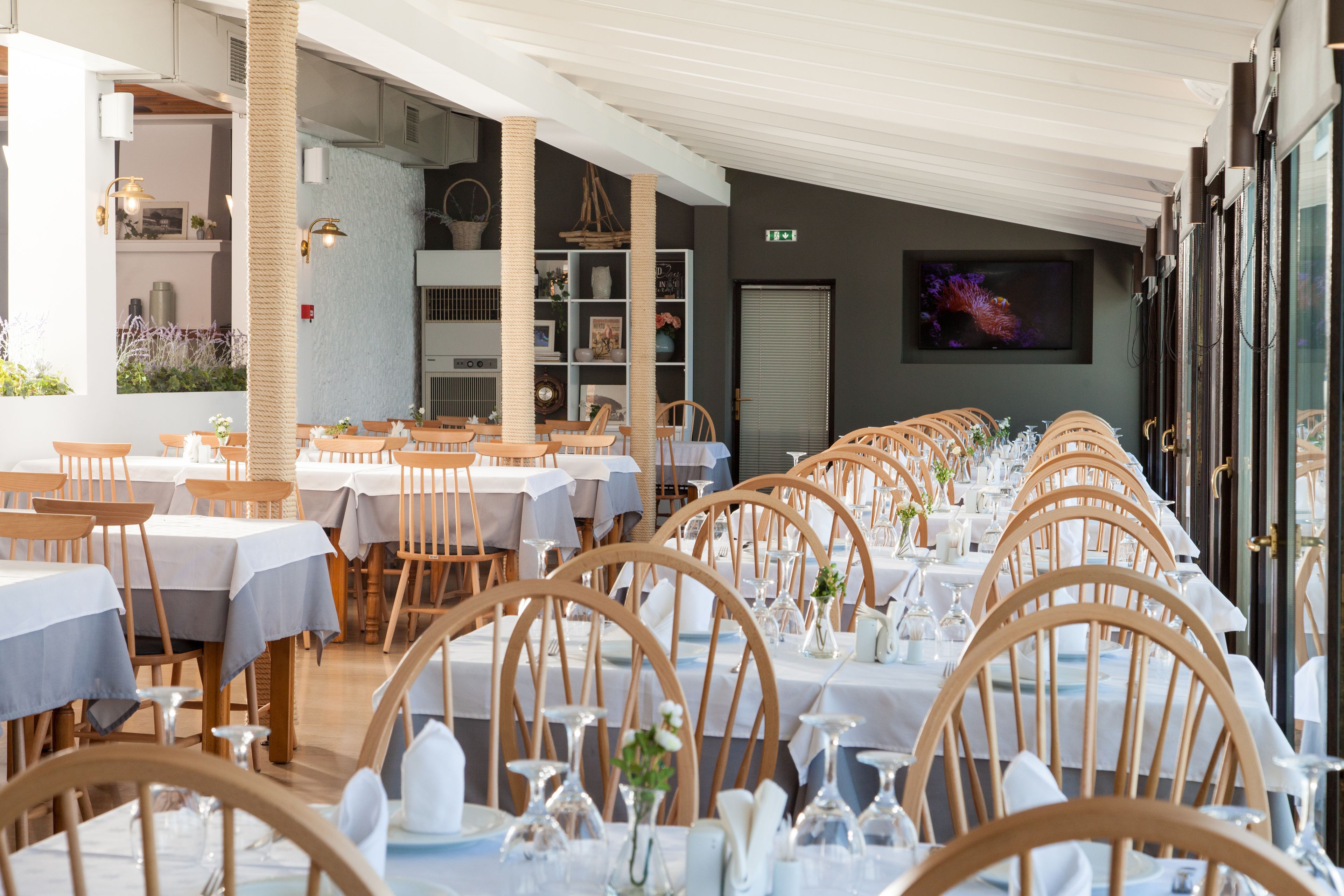 A restaurant with long tables, wooden chairs, and white tablecloths.