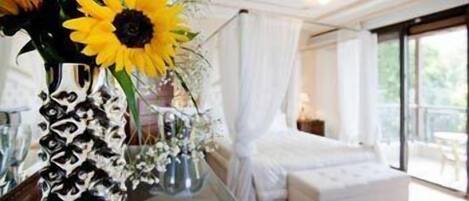 A room with a large window, a white bed, and a vase with sunflowers.
