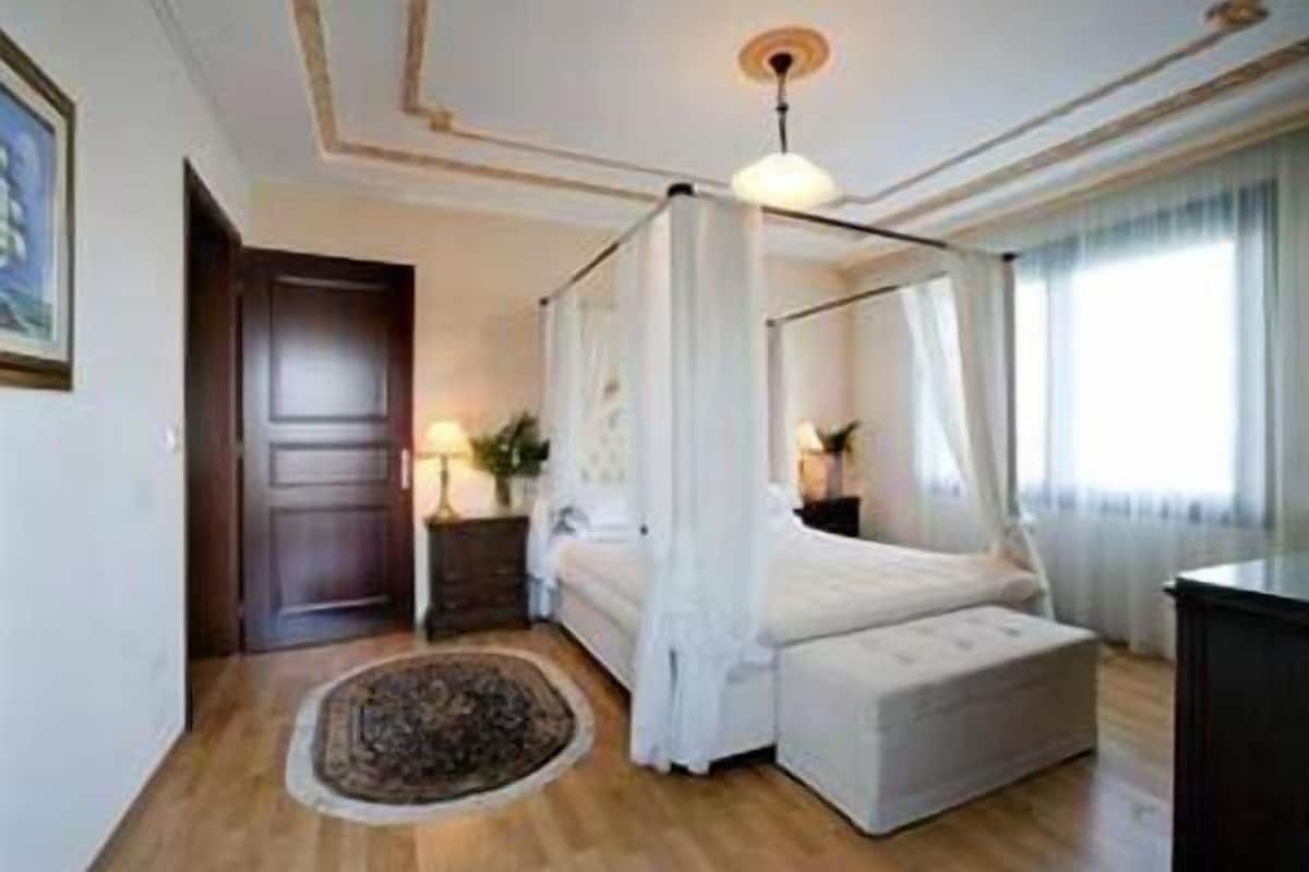 Una cama con dosel, una mesita de noche con lámpara, una alfombra y un amplio ventanal con cortinas.