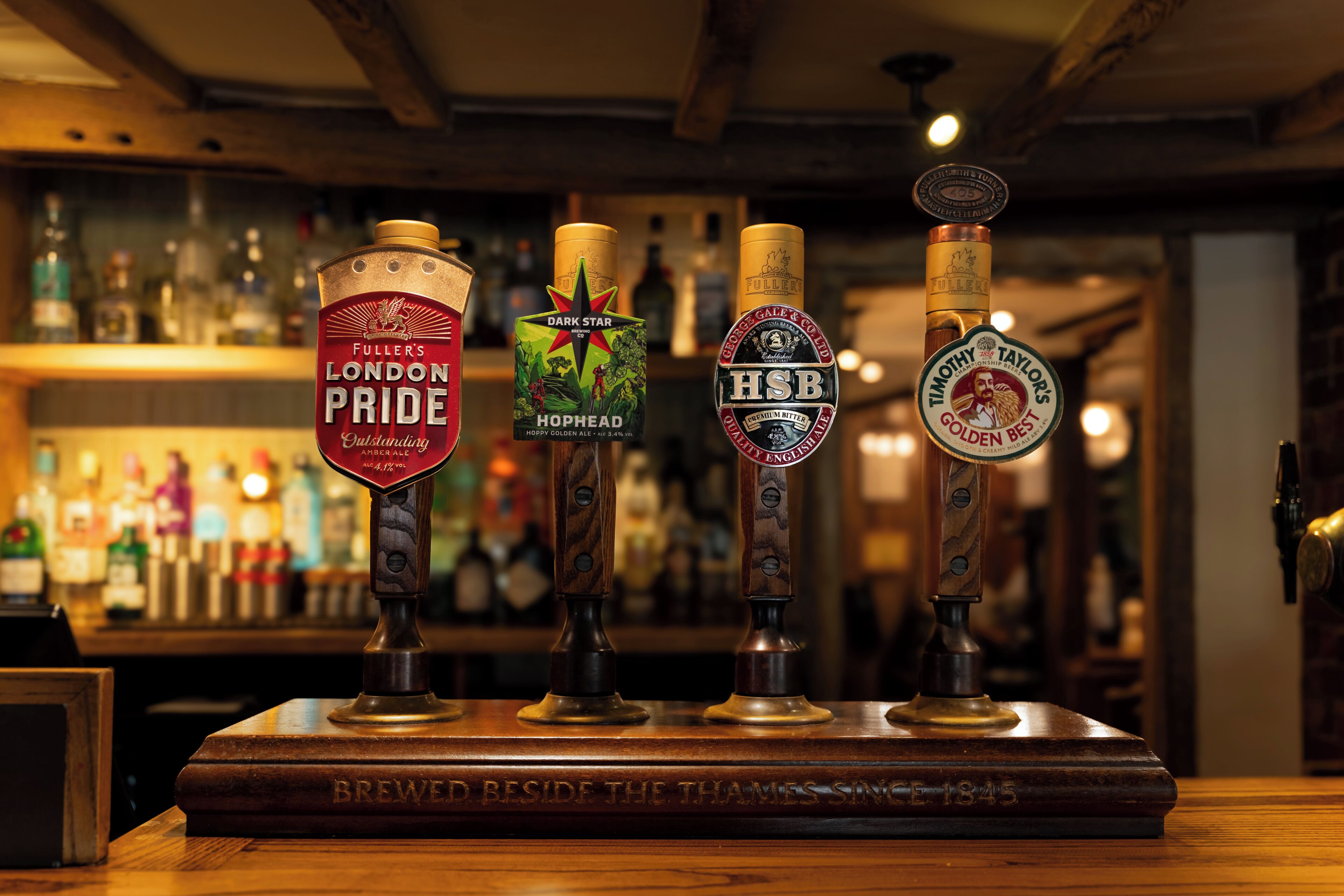 Quatro torneiras de cerveja numa bancada de bar de madeira, com um letreiro que diz 'BREWED BESIDE THE THAMES SING'.