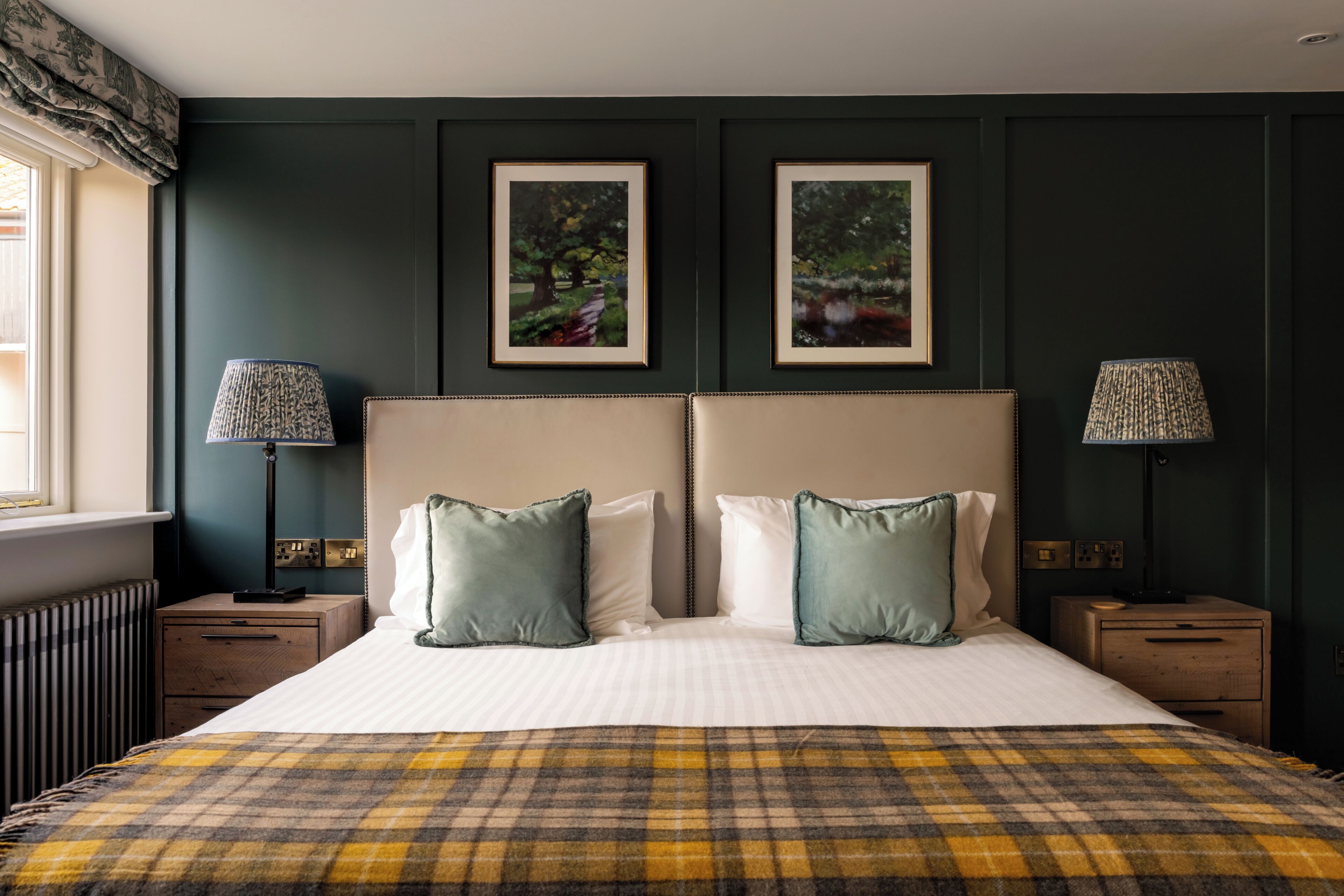 A bedroom with a large bed, two bedside tables, a window with curtains, and two framed pictures on the wall.