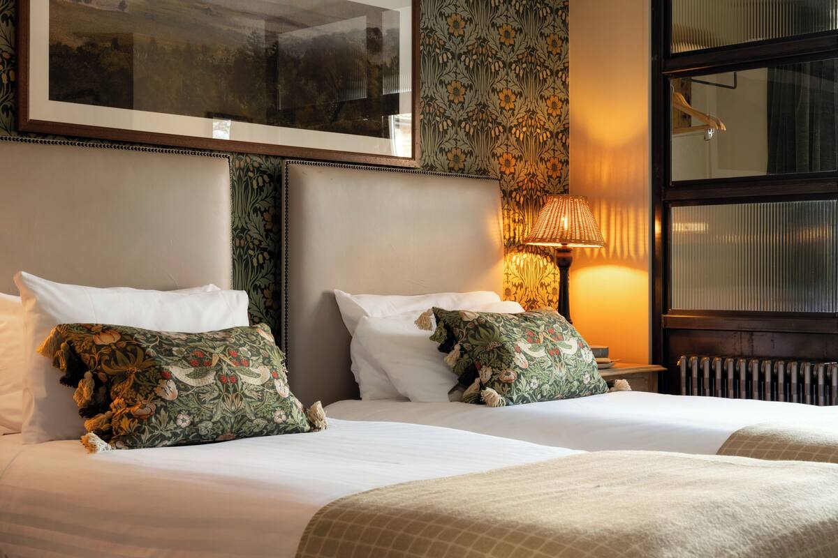 Une chambre d’hôtel avec deux lits, un tableau encadré au-dessus de la tête de lit, un papier peint à motifs et une lampe sur la table de chevet.