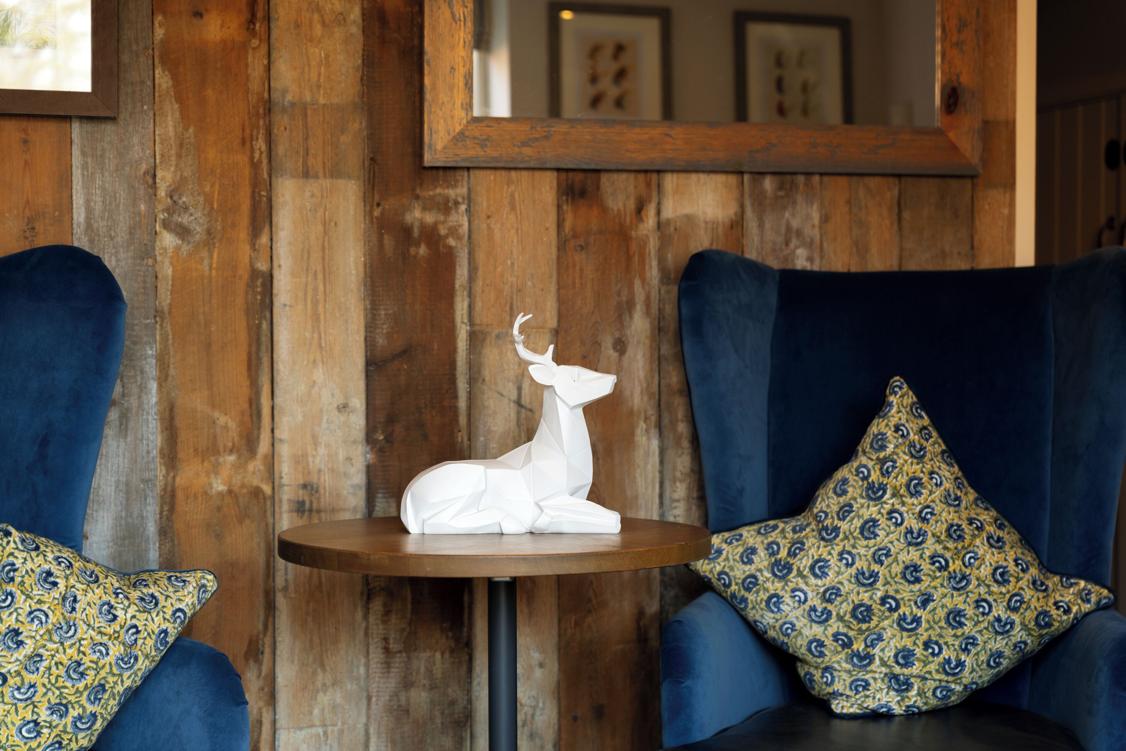 Une pièce aux murs en bois, avec une table ronde et deux fauteuils bleus ornés de coussins à motifs.