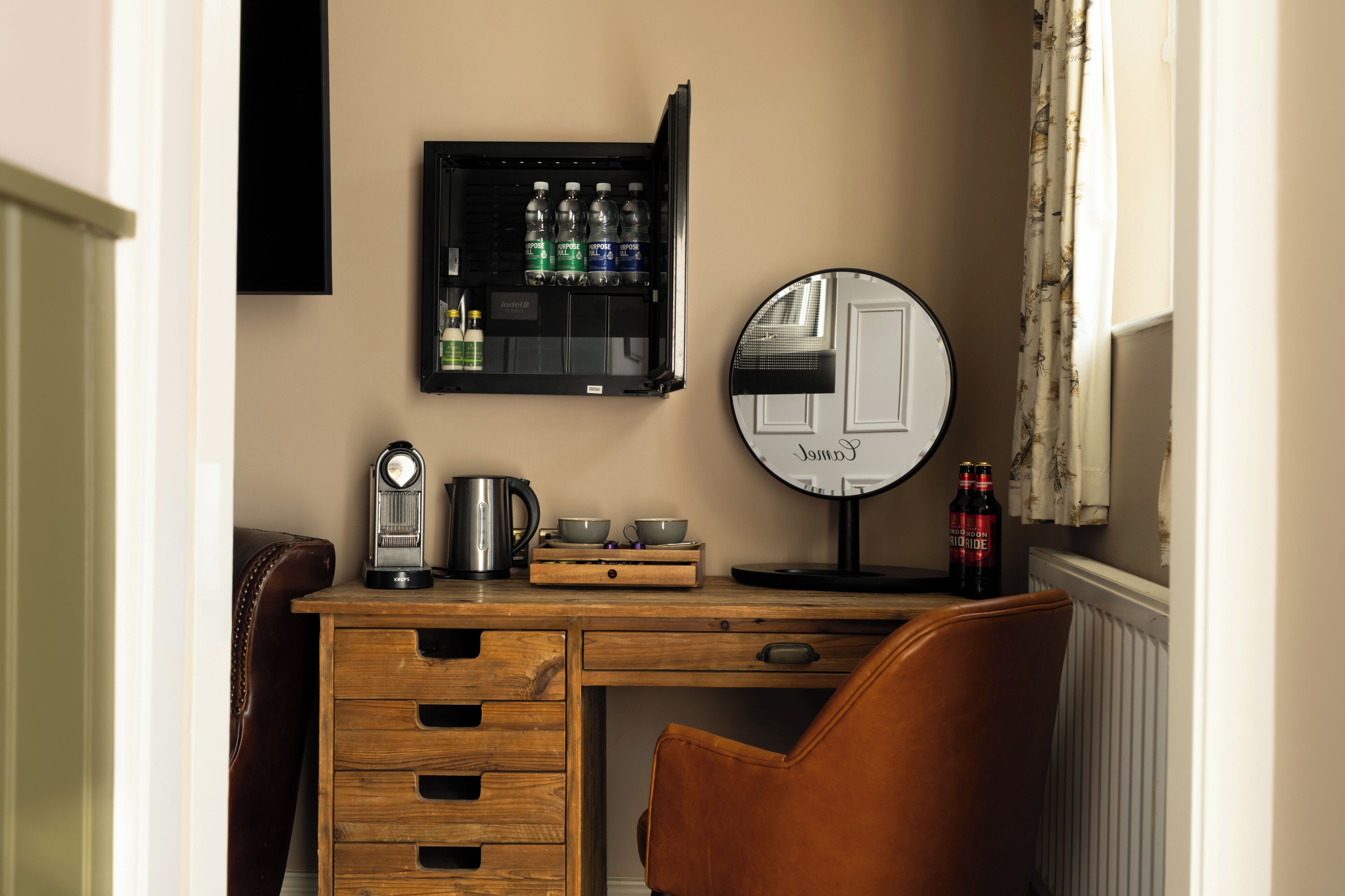 A hotel room with a wooden desk, a coffee maker, a kettle, a mirror, and a minibar with bottled water.