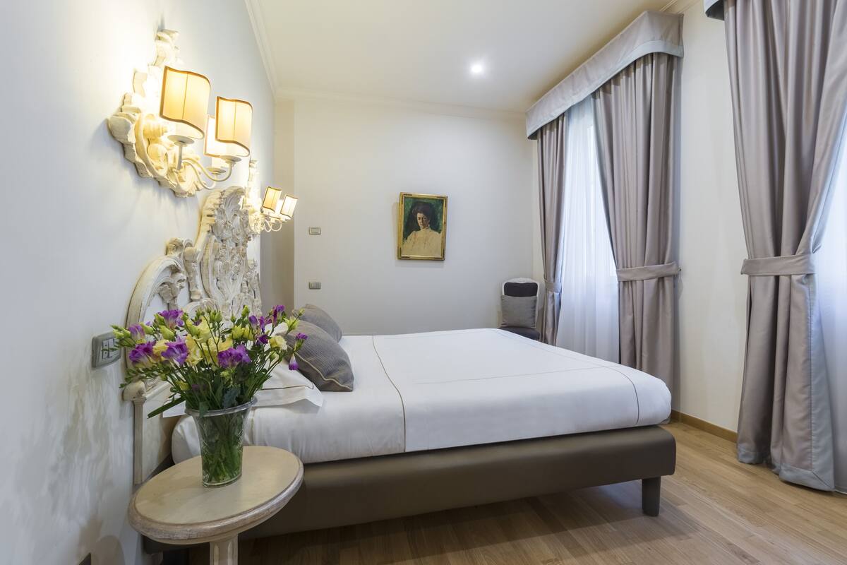 A bedroom with a bed, bedside table, wall-mounted lamp, and a vase with flowers.