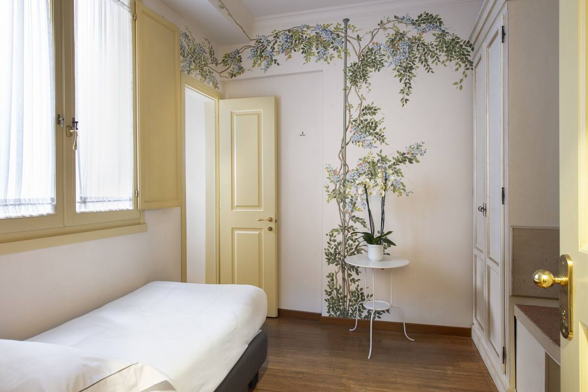 A bedroom with a bed, a window, a door, a plant, and a small table.