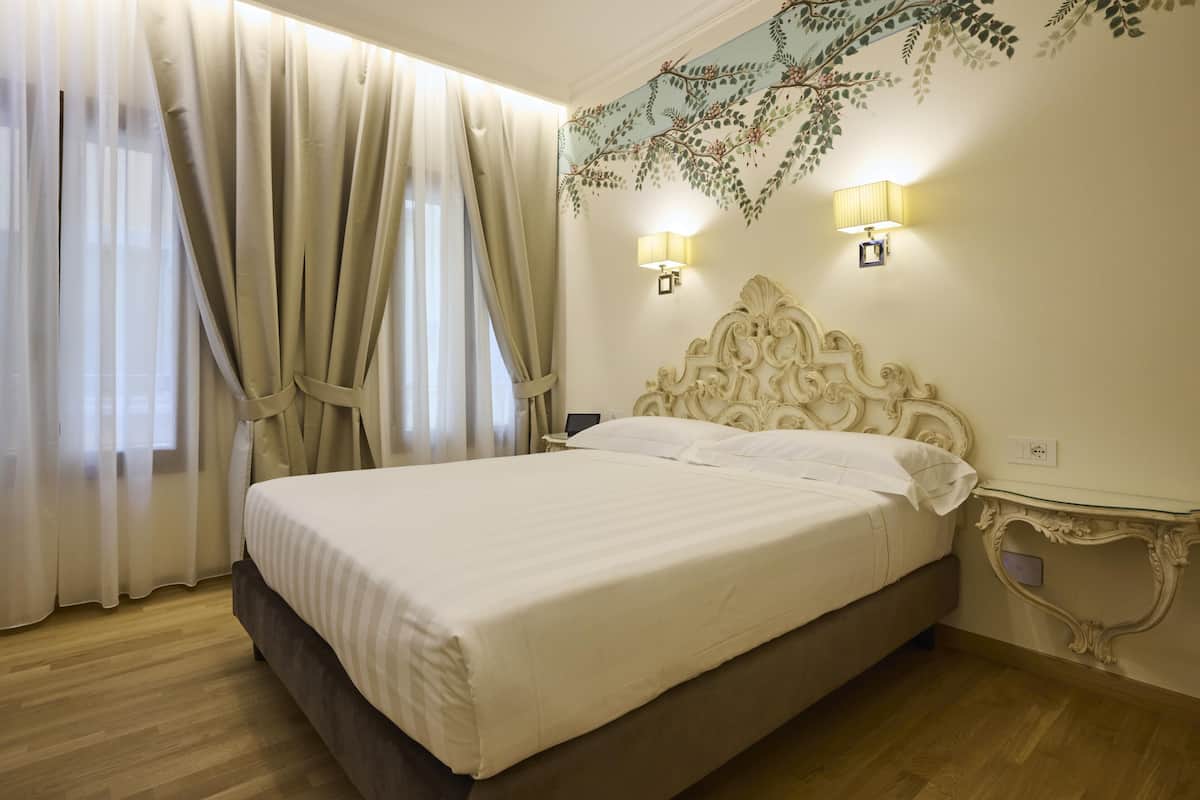 A neatly made bed in a hotel room with a large window, sheer curtains, and wall-mounted lamps.