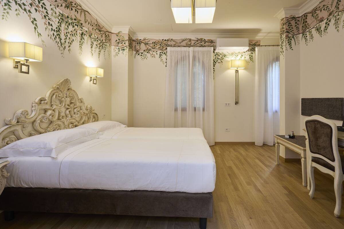 A bedroom with a large bed, a desk, and a chair. There are two lamps on the wall, and a window with curtains.