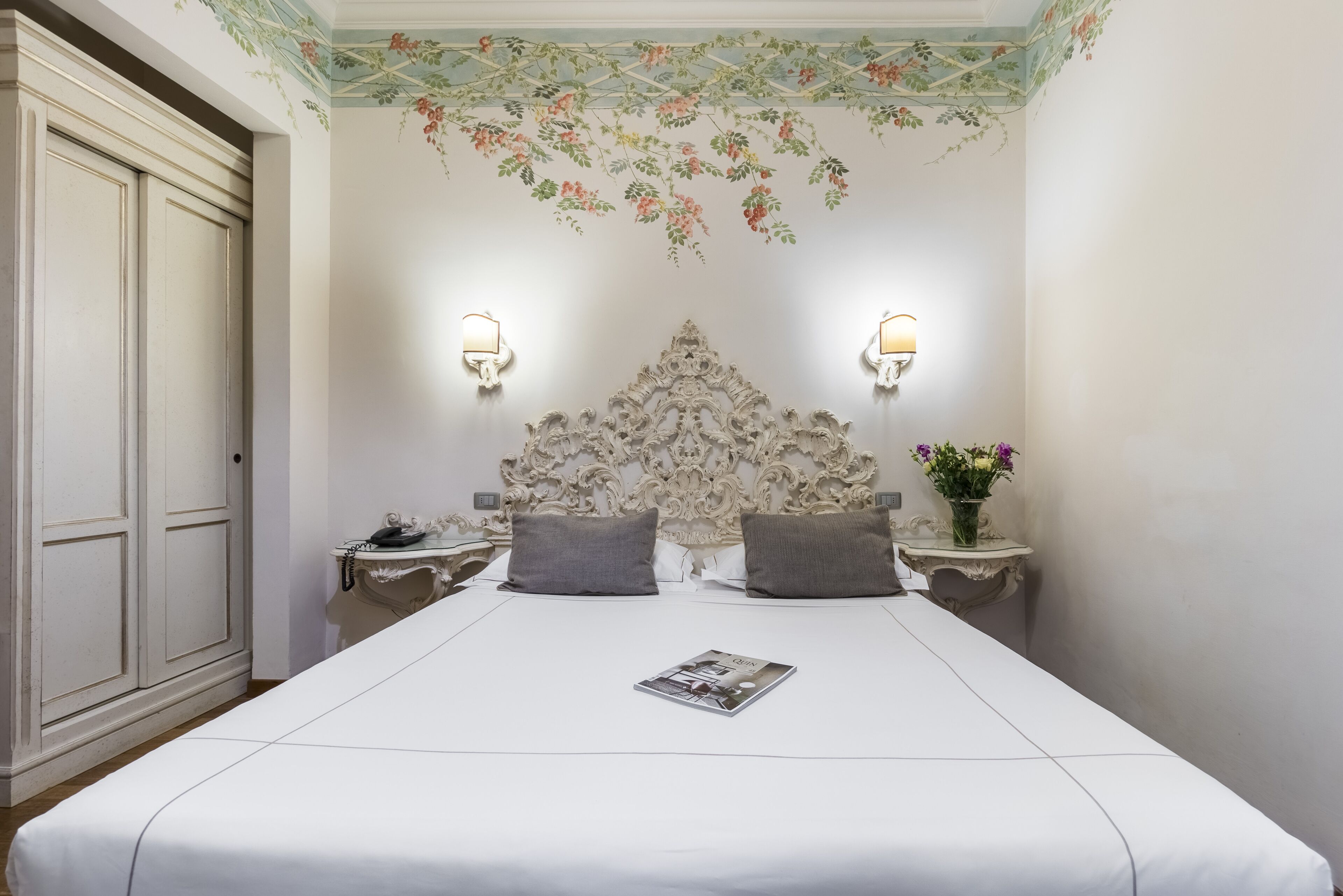 A hotel room with a large bed, two bedside tables with lamps, a decorative headboard, and floral wall art.