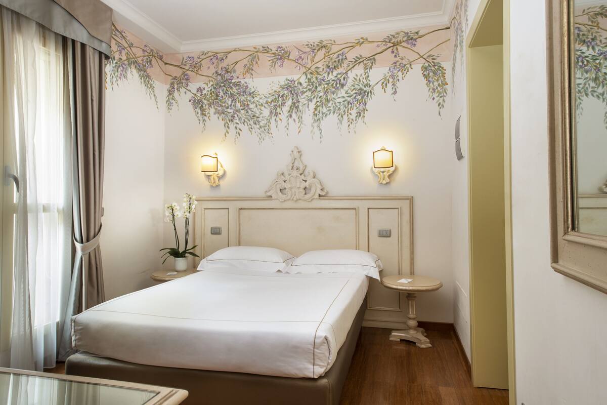 A hotel room with a large bed, two bedside tables, a nightstand, a mirror, and a decorative wall hanging.