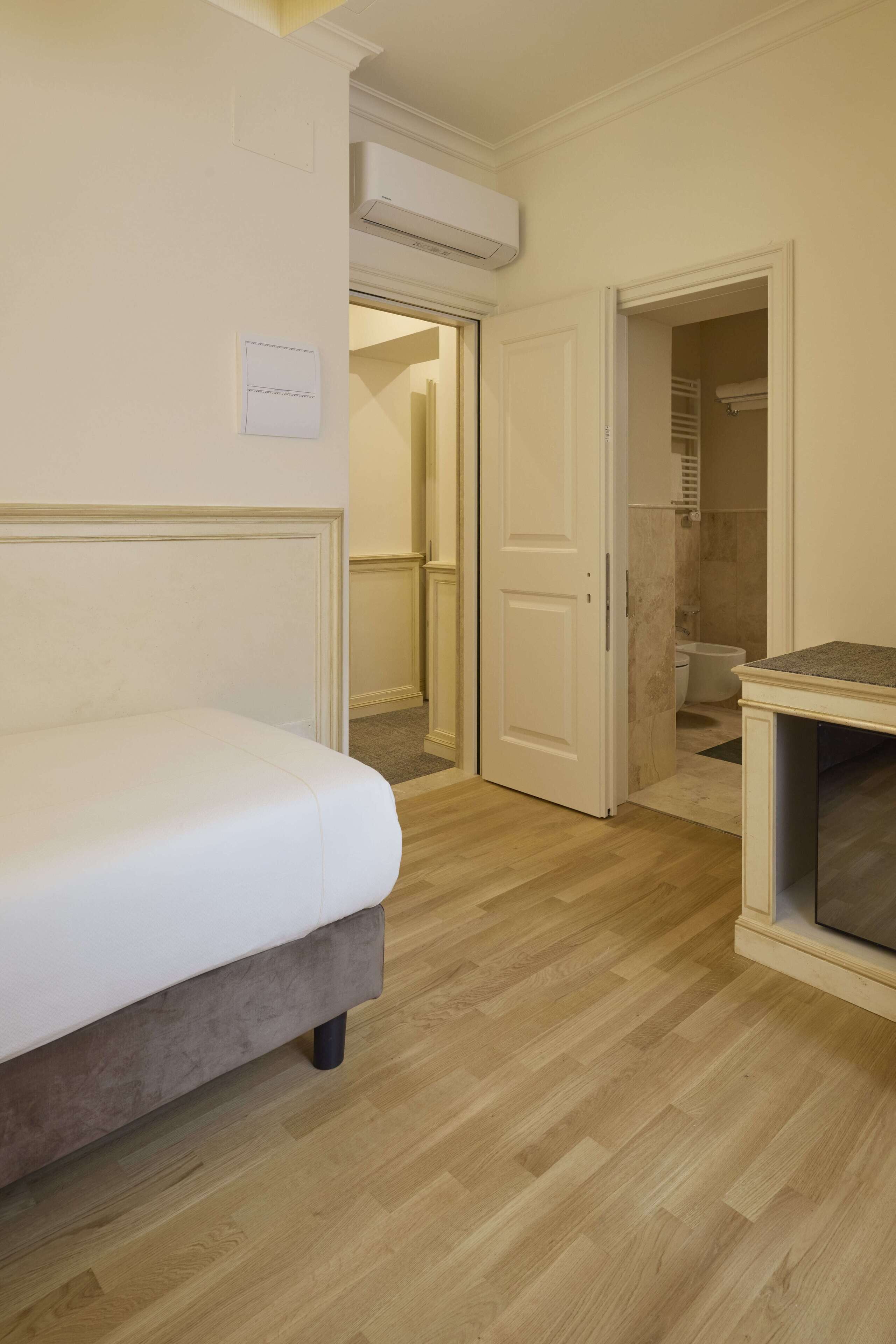 Une chambre à coucher avec un lit, un plancher en bois, un appareil de climatisation blanc et une salle de bain visible à travers une porte ouverte.