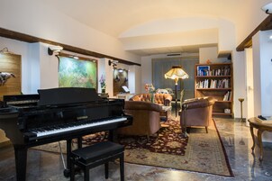 Un pianoforte a coda, divani in pelle, una libreria e un lampadario in un'ampia sala da soggiorno con pavimento in marmo e un grande quadro appeso al muro.