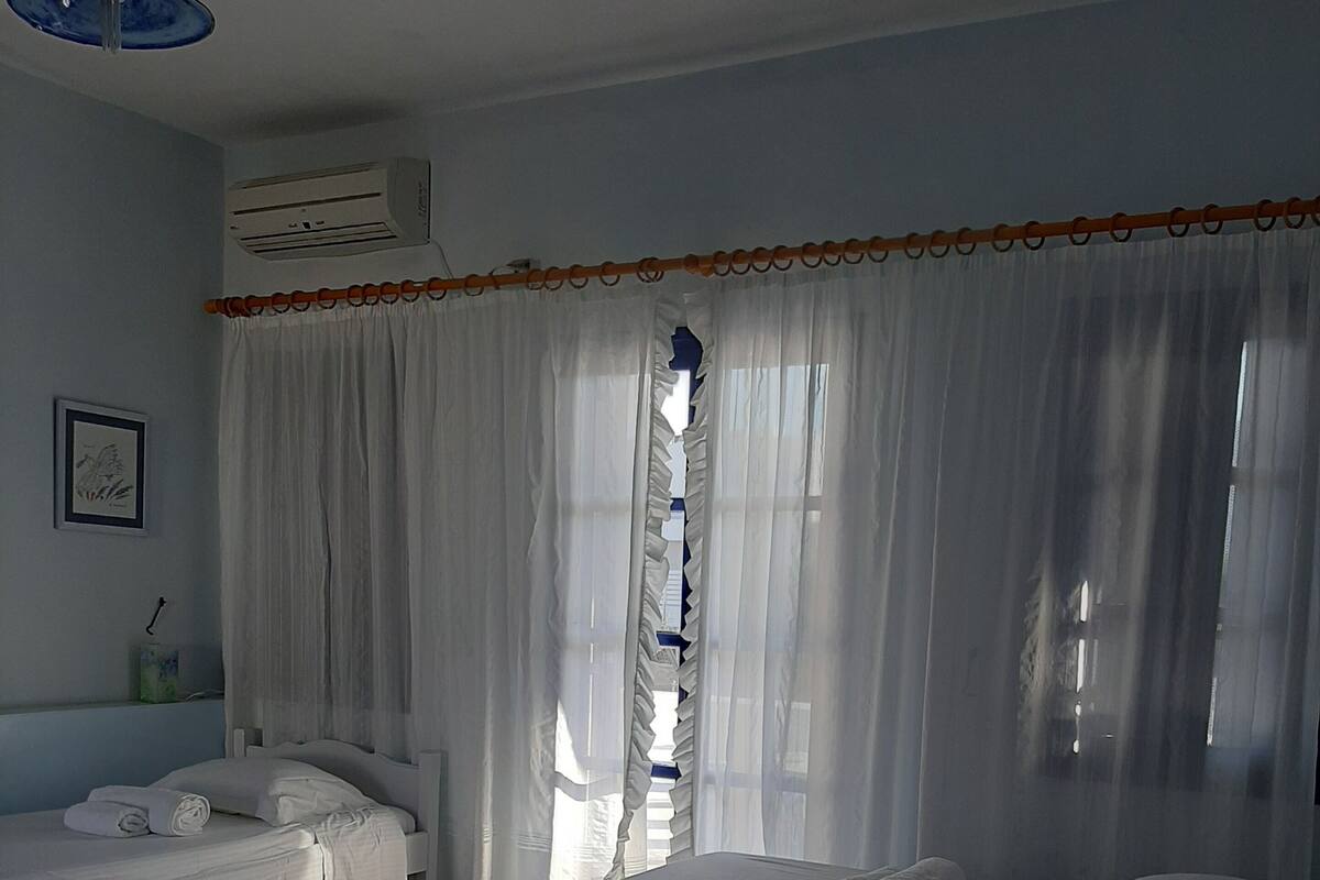 A hotel room with two beds, a ceiling fan, an air conditioning unit, and a window with sheer curtains.
