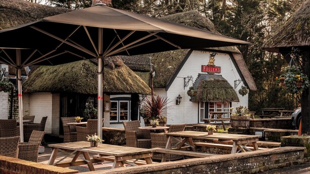 An outdoor seating area with wooden tables and wicker chairs, a thatched-roof building, and hanging flower baskets.