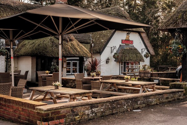 An outdoor seating area with wooden tables and wicker chairs, a thatched-roof building, and hanging flower baskets.