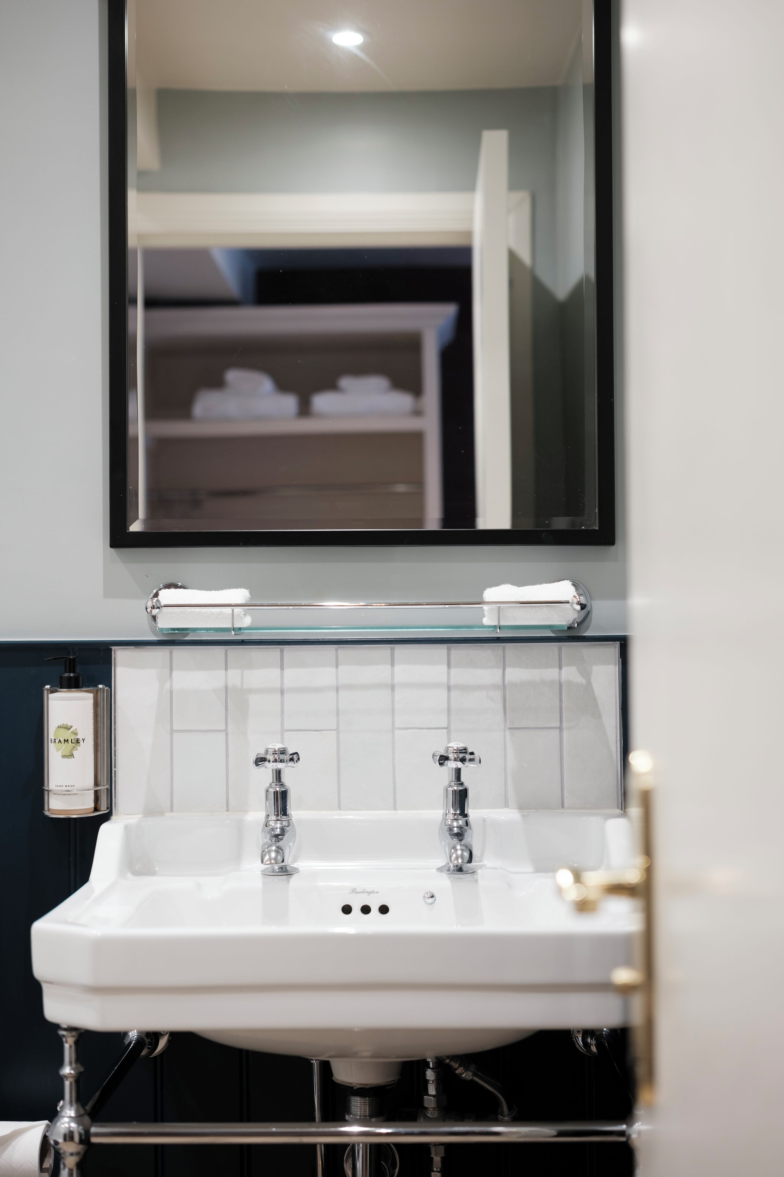 Baño con lavamanos blanco, dos grifos y un estante de vidrio sobre el lavamanos.