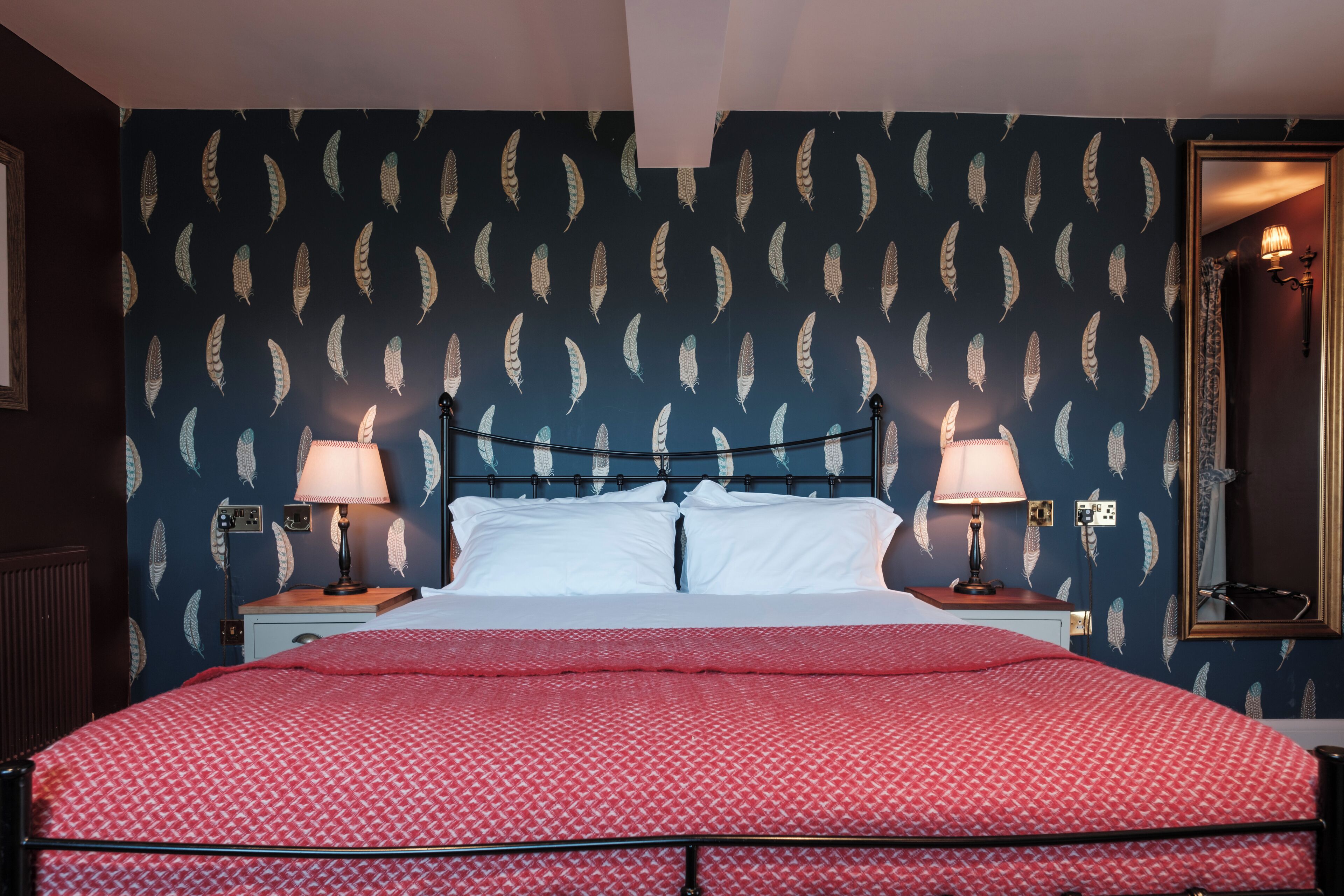 Un lit bien fait, recouvert d’une couvre-lit à motifs rouges, avec deux lampes de chevet et un mur orné d’un motif de plumes.
