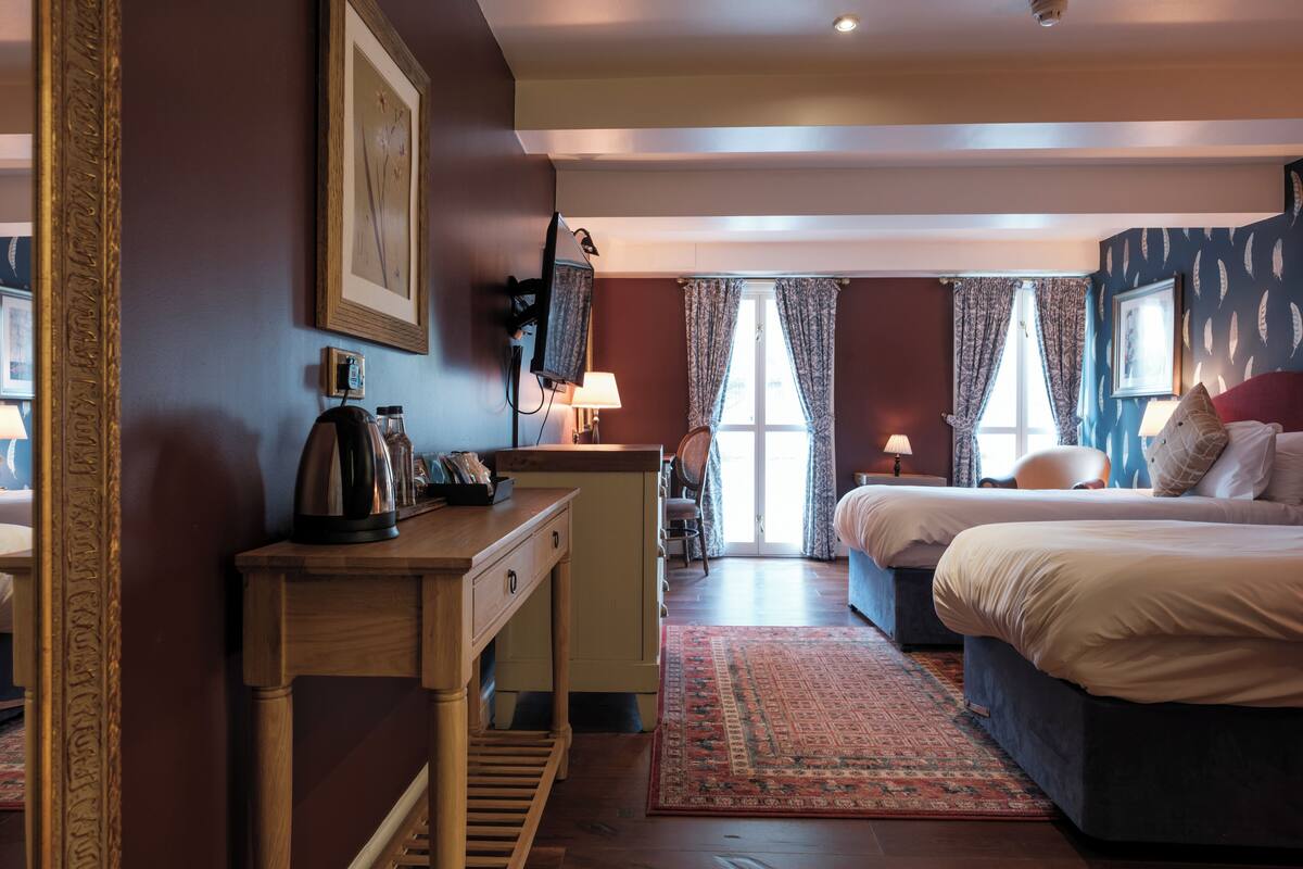 Quarto de hotel com cama, escrivaninha, cadeira, luminária e janela com cortinas.